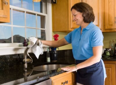 Cheerful Maid Polishing Kitchen Sink Faucet Cheerful maid polishing a brushed nickel faucet in a high end luxury kitchen. Cleaning woman has a smile with eyes focused on the task at hand.